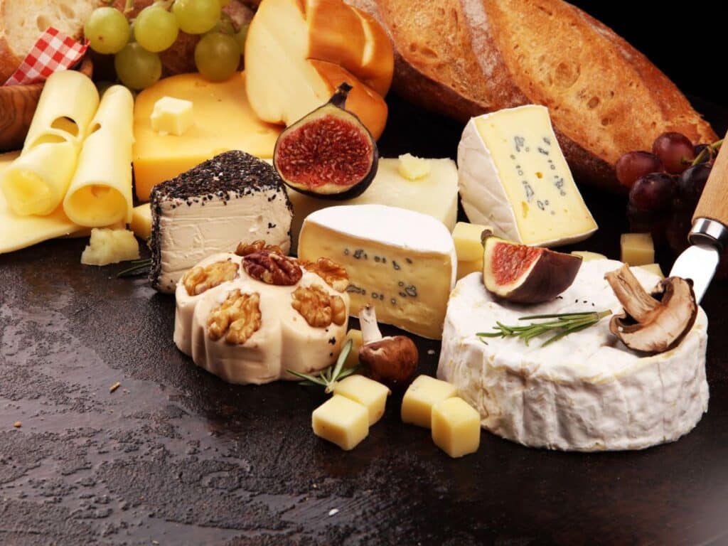 A rustic cheese board featuring a selection of the best French cheeses, including creamy Brie, blue-veined Roquefort, and nut-topped soft cheese, surrounded by fresh figs, mushrooms, crusty baguette slices, and grapes. Cubes and slices of cheese are artfully arranged on a dark wooden surface for an inviting gourmet presentation.