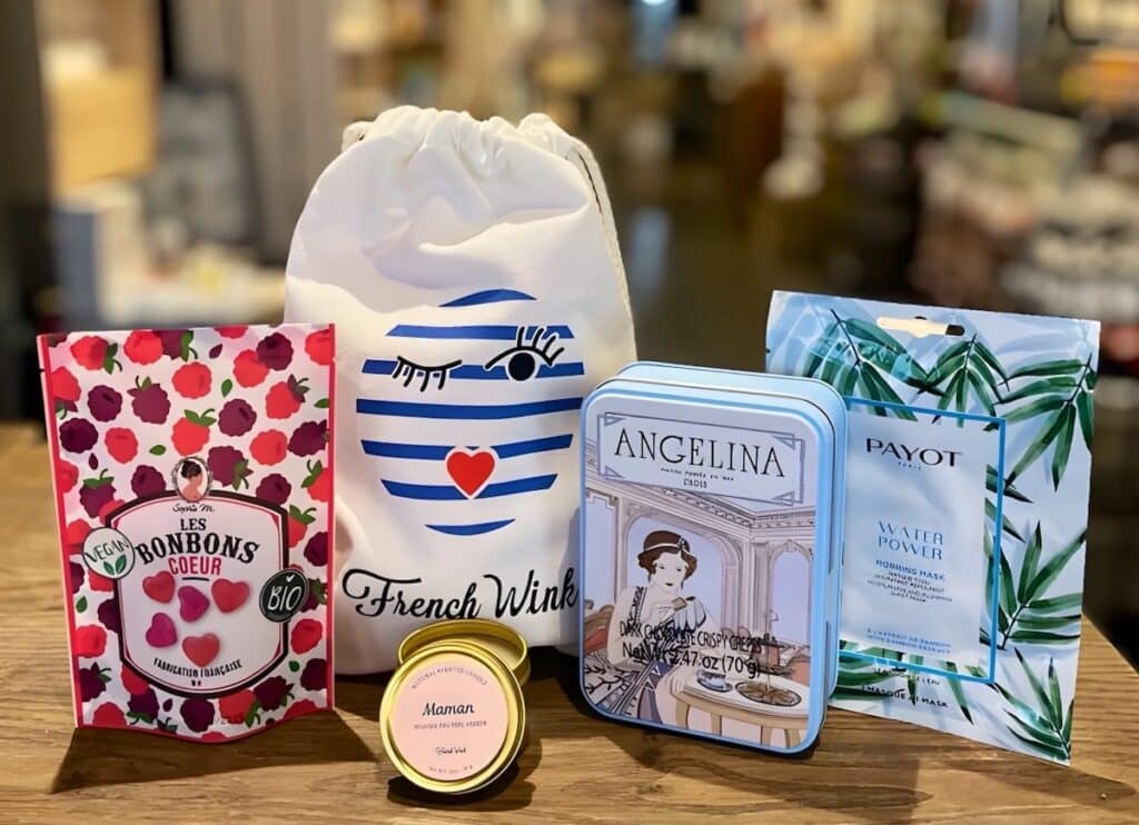 A curated French gift set displayed on a wooden table, featuring organic heart-shaped vegan candies, a "Maman" scented candle, a French Wink drawstring bag, Angelina dark chocolate crisps in a decorative tin, and a Payot hydrating face mask. A thoughtful and stylish selection of French gifts for Christmas with a mix of sweet, self-care, and artisanal items.