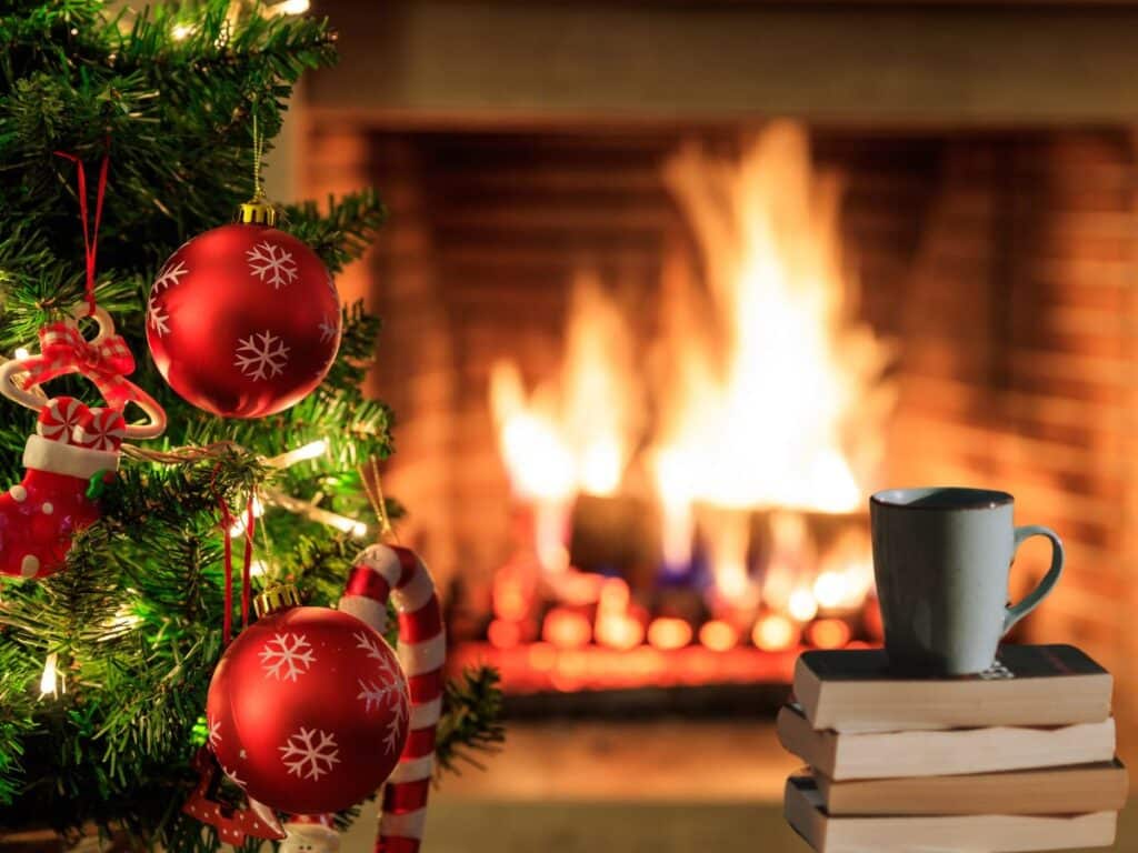 A cozy holiday scene with a decorated Christmas tree featuring red ornaments and candy canes, a stack of books, a mug, and a roaring fire in the background. This warm setup evokes the perfect setting to read books set in France at Christmas.