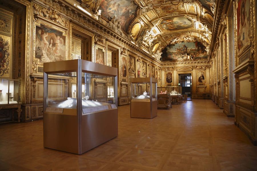 Lavishly decorated Louvre gallery lined with gilded frames and ornate ceiling frescoes, displaying royal jewels in glass cases under soft lighting. The scene sets the stage for a possible Heist at the Louvre.