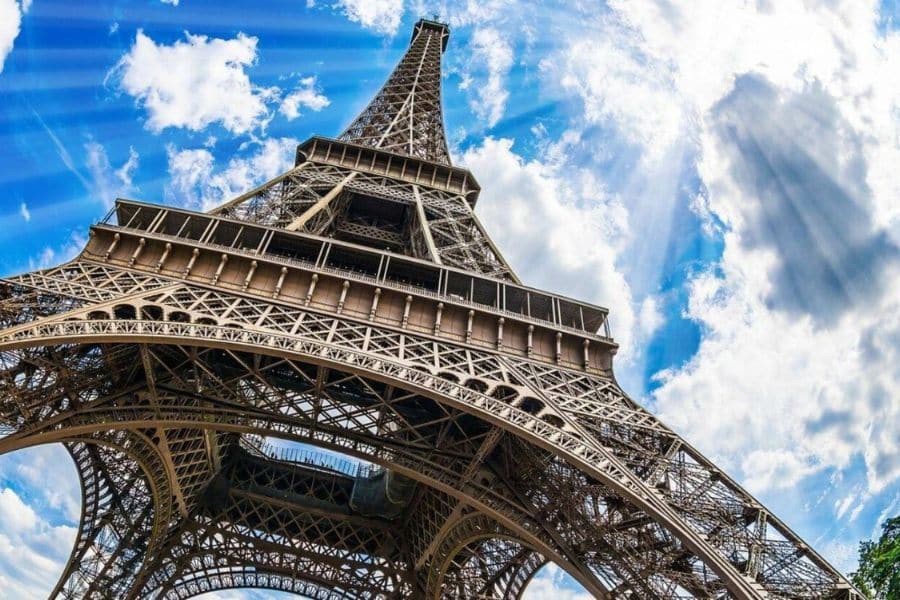 Dramatic upward view of the Eiffel Tower against a vibrant blue sky with sunbeams and clouds. The image emphasizes the tower’s intricate iron lattice structure, subtly referencing where the names inscribed on the Eiffel Tower are located beneath the first platform.