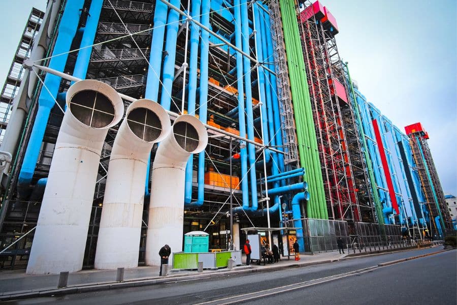A street-level view of the Centre Pompidou in Paris, showcasing its iconic exposed pipes in bold colors like blue, red, and green, with large white air ducts in the foreground.