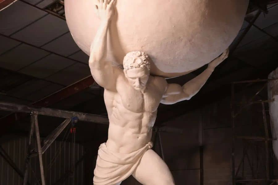 Close-up of a colossal white statue of Atlas straining under the weight of a huge globe, set in a warehouse with scaffolding. The powerful physique and determined expression embody the essence of a mythological Guardian of Liberty bearing the world’s weight.