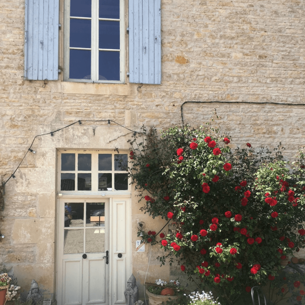 The façade of a charming stone house in the Charente region, featuring traditional architecture with a natural stone finish. A light-colored door is flanked by two statues, with a glass panel above allowing light into the entrance. To the right, a lush rose bush with vibrant red blooms adds a splash of color against the neutral tones of the building. Above, a window with pale blue shutters underlines the rustic French countryside aesthetic.