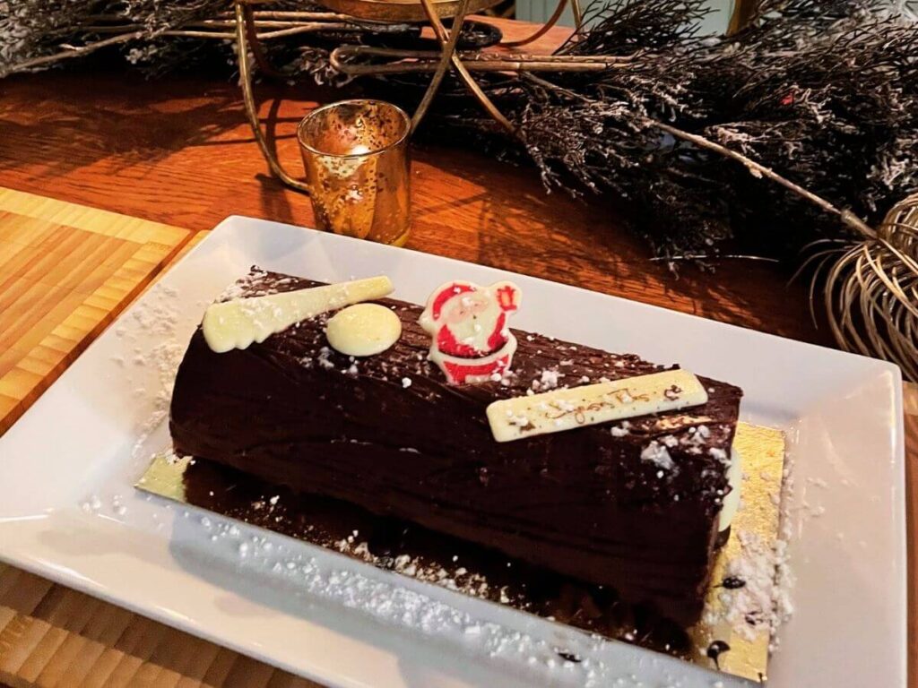 A Christmas chocolate log sat on a white plate and decorated with icing sugar with a little santa on the top