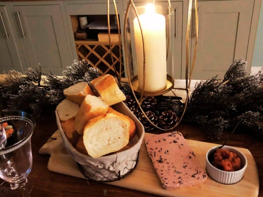 A basket of freshly cut French bread sat on a wooden platter with a big slab of pate and some pickles with a bit lit candle behind