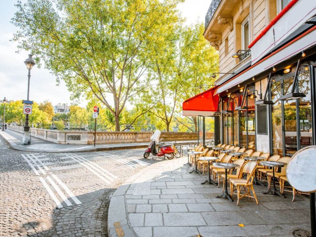 The facade of a cafe in Paris with a red awning and tables and chairs set up on the pavement with green trees across the street