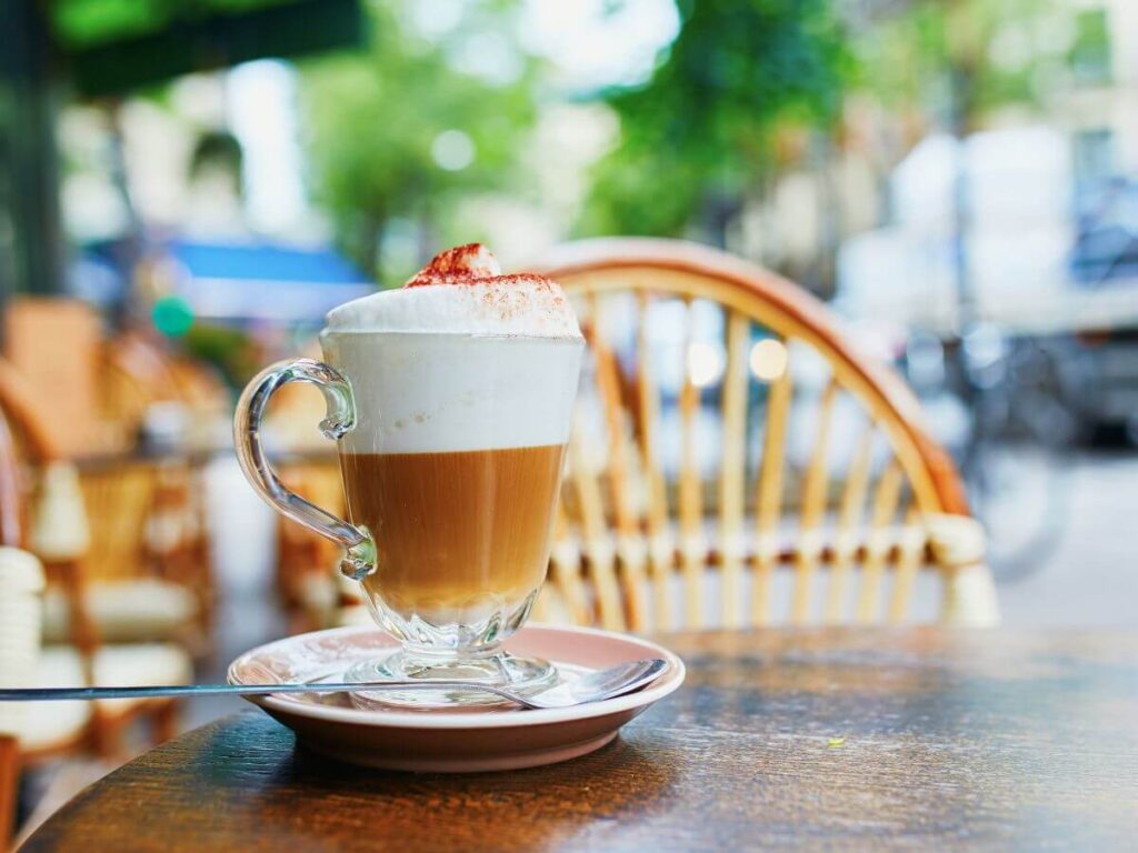 A coffee in a glass with lots of cream on top sat on a wooded table with a chair behind it