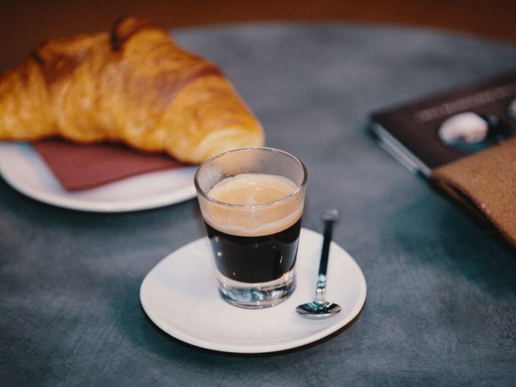 A small espresso in a glass on a white saucer on a table with a croissant on a white plate