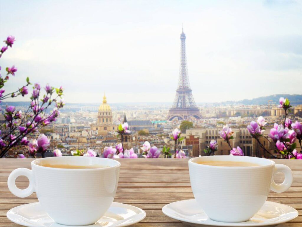 Two white coffee cups on saucers on a wooded table with a distant view of the Eiffel Tower in Paris and some pink flower bushes in front