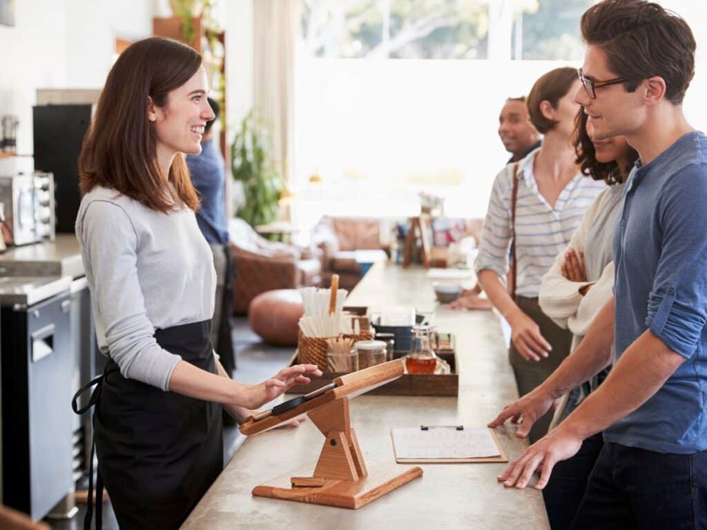A cheerful barista in a casual sweater and apron engages with customers at a modern, sunlit coffee shop counter, with an iPad register and a menu in view, encapsulating a daily slice of French life and the culture of greeting