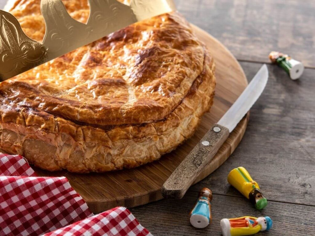 A pastry dish on a wooden platter with a knife by the side, a red and white French napkin, some small Christmas ornaments and part of a gold crown sat on the edge of the pastry