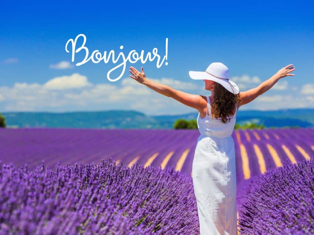 A woman in a white sundress and wide-brimmed hat extends her arms joyfully among vibrant lavender fields under a clear blue sky, with the word 'Bonjour!' written above in elegant script, symbolizing a warm greeting or hello in French.