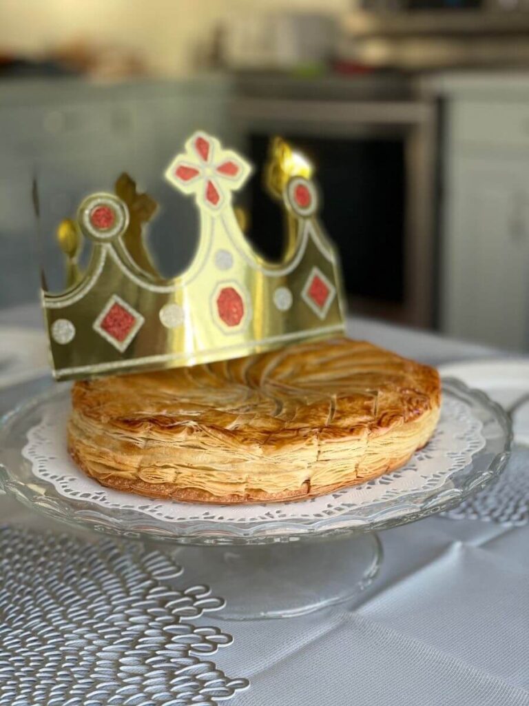 A pastry based cake sat on a doilie on a glass plate with a toy gold crown sat on top of it.