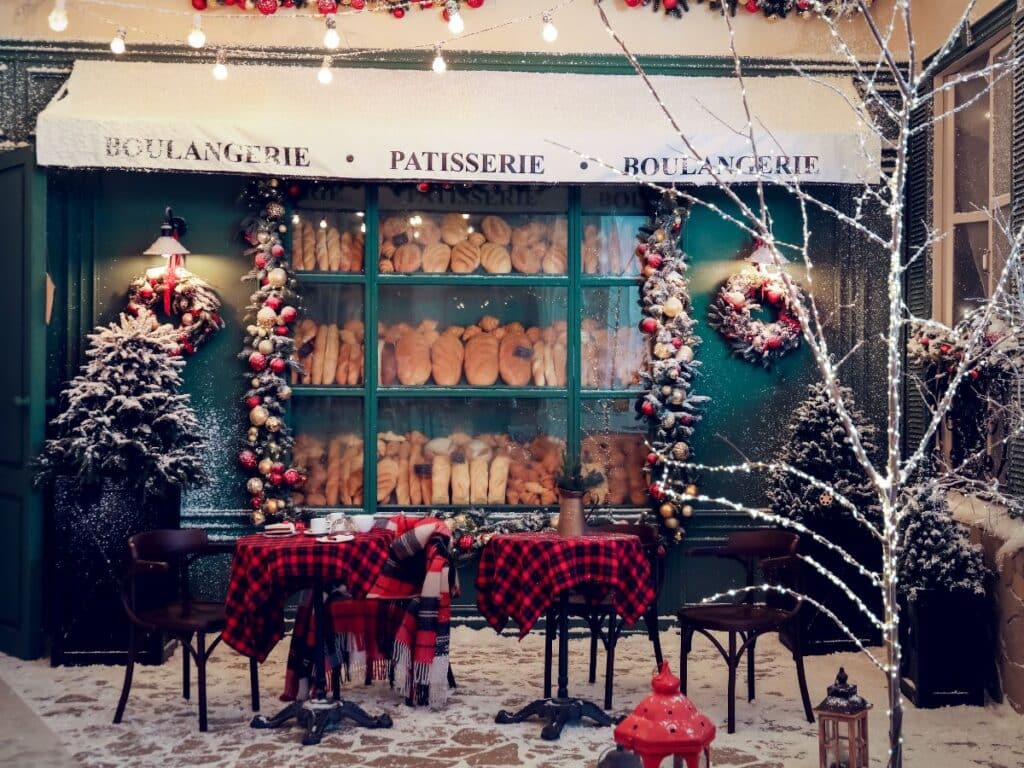 A festive French bakery facade adorned with Christmas wreaths and lights, dusted with fresh snow. The display window showcases an array of bread and pastries, while a charming outdoor seating area with red plaid blankets invites holiday cheer.