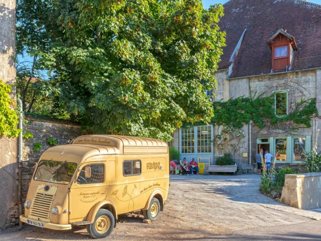 An old-fashioned cream-colored delivery van with "Anis de Flavigny" branding parked in a serene street of Flavigny-sur-Ozerain, France. Behind the van is a stone building with greenery around the windows and a sign that reads "Anis de Flavigny." The warm light of the setting sun enhances the quaint charm of the scene. People are seated casually on a bench by the building, enjoying the peaceful ambiance of this picturesque French village.