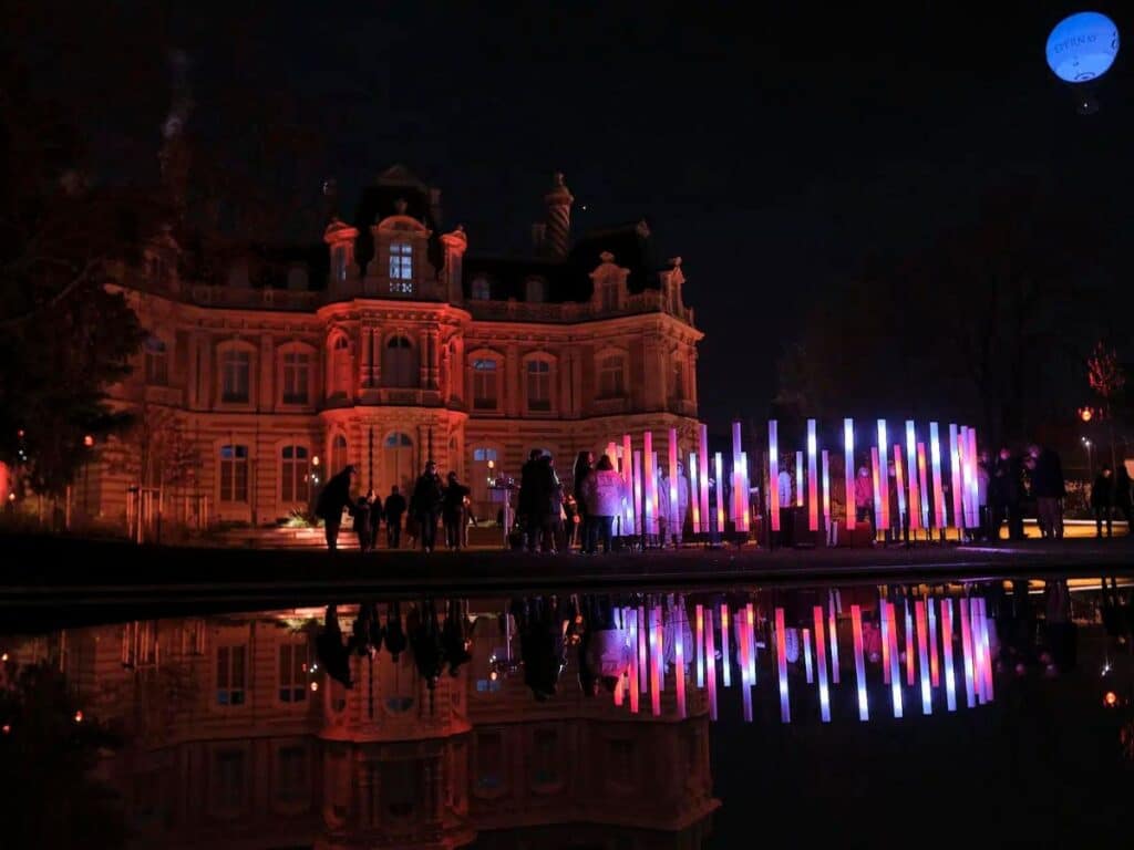 Visitors gather at night in front of an illuminated historical building during Les Habits de Lumière festival in Epernay, with vibrant neon light installations reflected in a still pond.