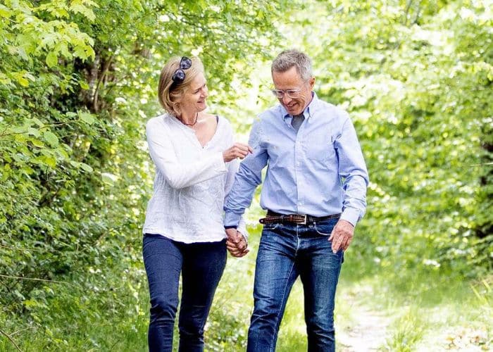 A middle-aged couple, casually dressed, enjoys a leisurely walk together along a lush, green woodland path. The woman, wearing a white blouse and sunglasses pushed back on her head, looks at the man with a smile, gesturing with her hand. The man, in a light blue shirt and jeans, returns the smile, looking down slightly as they hold hands. The natural sunlight filters through the canopy of leaves above them, highlighting the intimacy and contentment of a simple moment shared.