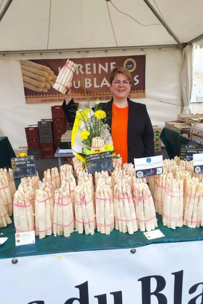 A woman in a black jacket and orange shirt stands happily behind a market stall at a food festival, holding a bundle of white asparagus. The stall is stocked with numerous bundles of white asparagus, clearly labeled with prices, and there's a banner in the background promoting "Reine du Blayais," suggesting a specialty in this product. A small floral arrangement adds a pop of color to the cheerful scene.
