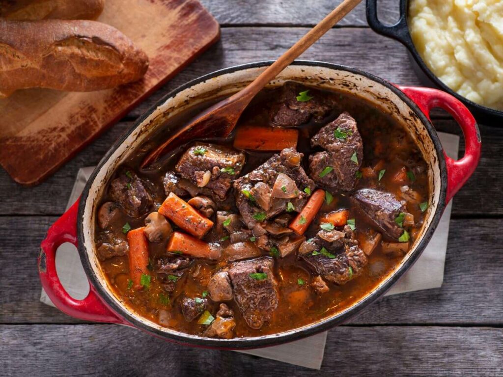 A hearty serving of Boeuf Bourguignon in a red cast-iron pot, accompanied by a side of creamy mashed potatoes and a crusty baguette. The stew is rich with tender chunks of beef, carrots, and mushrooms, garnished with fresh herbs. This traditional French dish is presented on a rustic wooden table, embodying the essence of French cuisine.