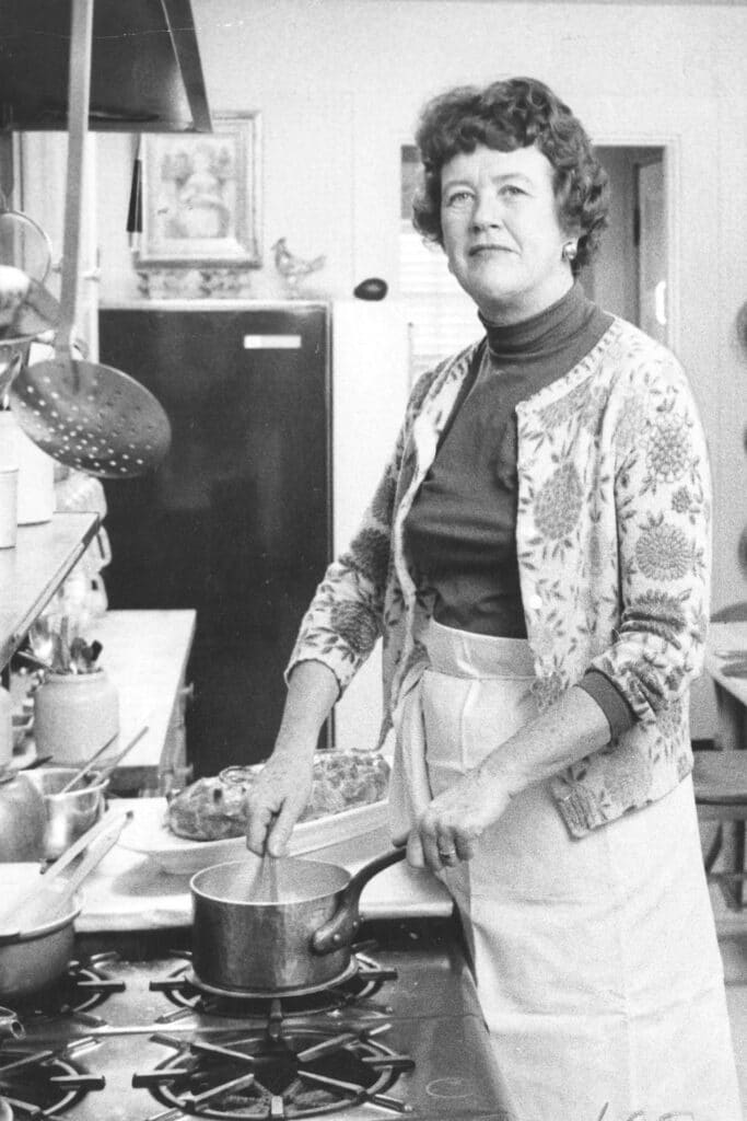 A black and white photo of Julia Child cooking in a kitchen, stirring a pot on the stove with a thoughtful expression, surrounded by various kitchen utensils and cookware.