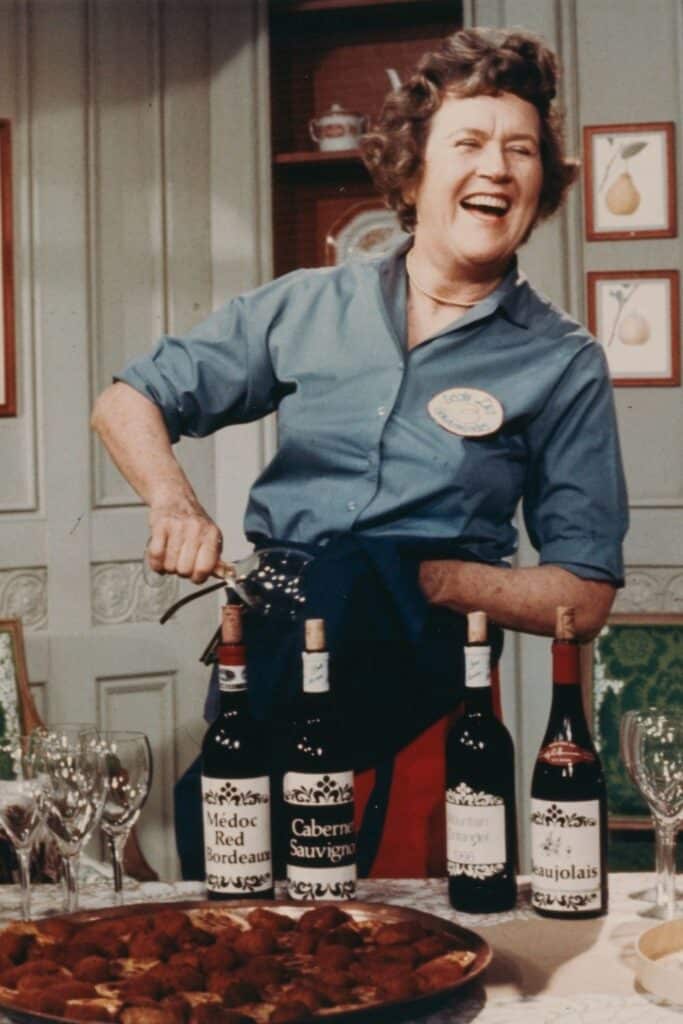 Colorful image of Julia Child laughing while opening a bottle of wine, with multiple wine bottles and a platter of food on the table, in a kitchen setting adorned with decorative wall plates.