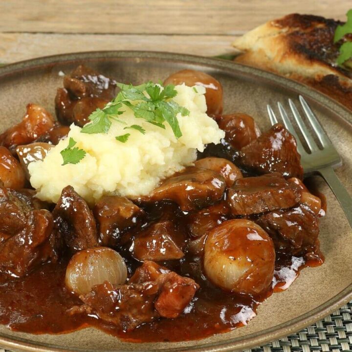 A plate of Beef Bourguignon served with a generous portion of creamy mashed potatoes, garnished with fresh parsley. The rich, dark sauce of the stew is glossy and coats the tender pieces of beef and pearl onions. Accompanied by a slice of grilled bread, this dish is a classic representation of French comfort food, ready to be enjoyed.