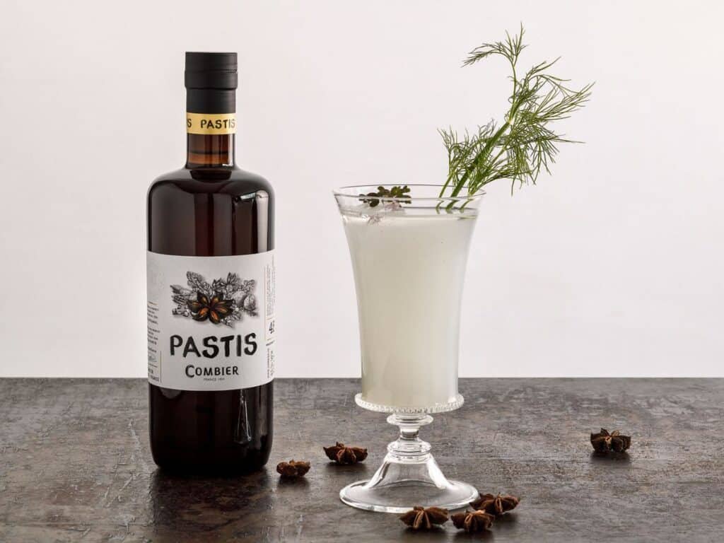 A bottle of Pastis Combier with a vintage-style label stands next to a glass of milky pastis garnished with a sprig of dill, presented on a dark, textured surface with scattered star anise pods around.