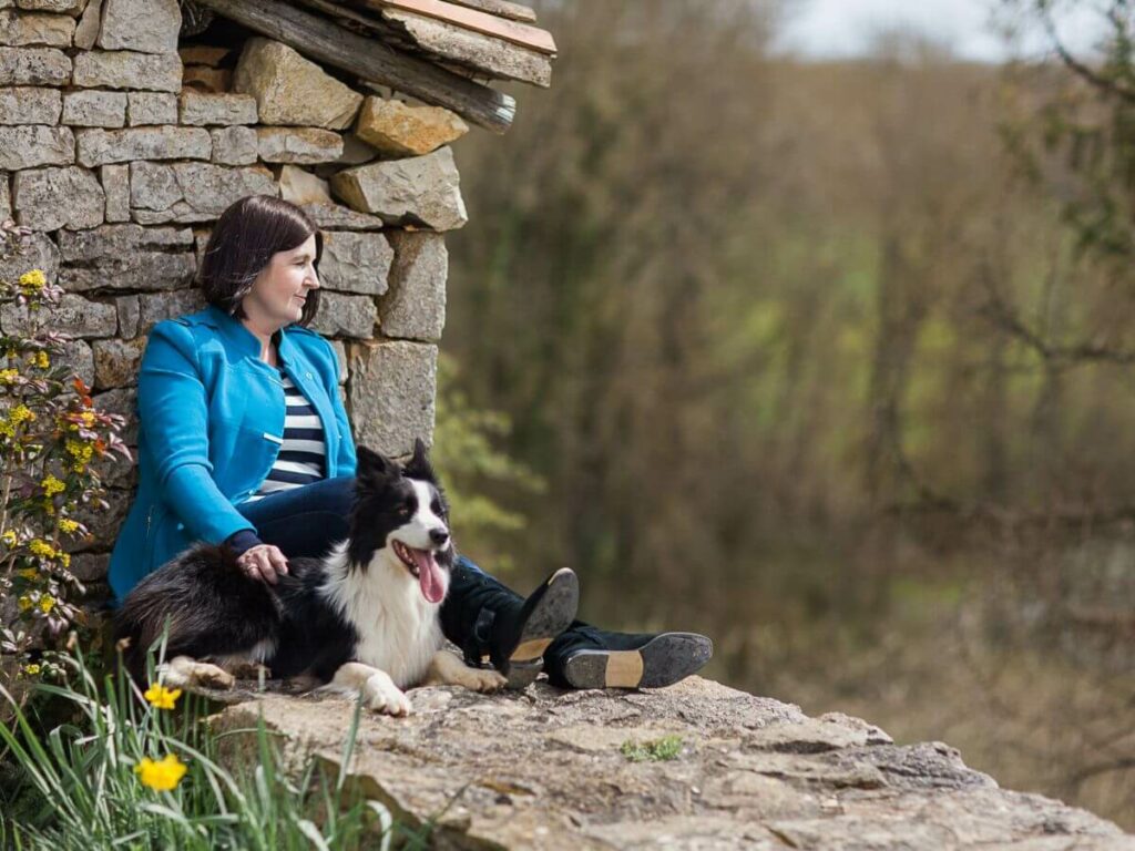 A woman sat on a stone wall in a blue coat with her dog in France