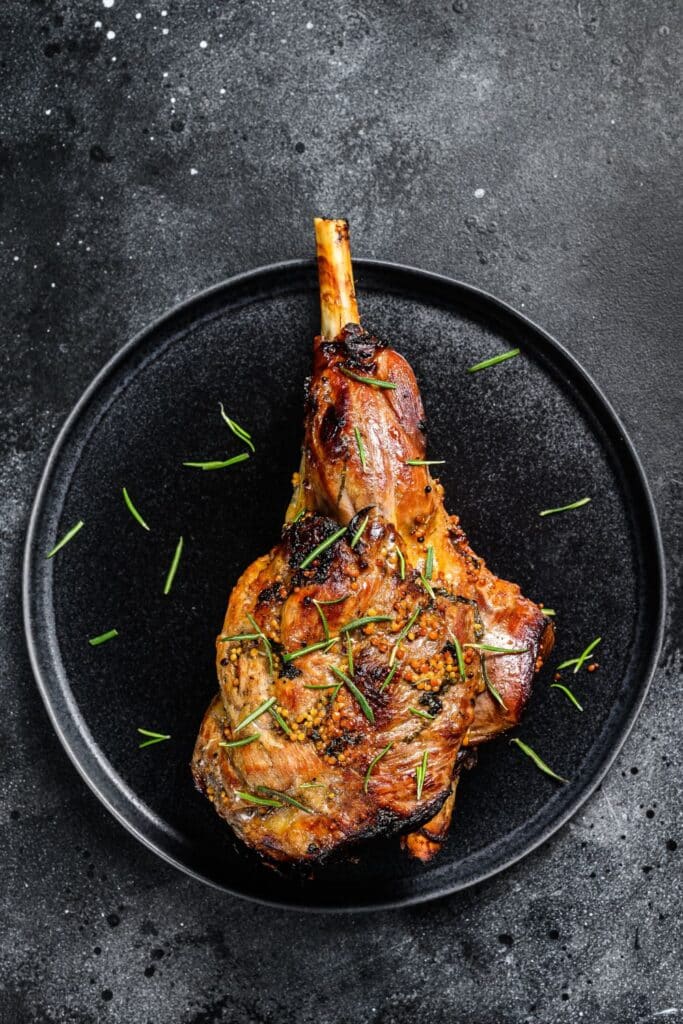 Succulent roasted leg of lamb with a caramelized exterior, garnished with rosemary on a matte black plate. The plate sits on a textured dark background, speckled with salt, highlighting the rich, savory dish often enjoyed as part of a traditional Easter meal in France.