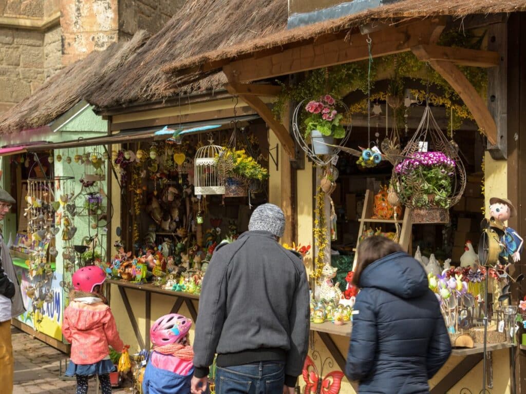 A lively Easter market scene in France with families browsing a stall adorned with colorful decorations, including floral arrangements, Easter figurines, and hanging ornaments. A thatched roof adds a rustic charm to the stall, which is nestled against the backdrop of an ancient stone building, capturing the festive spirit and cultural traditions.