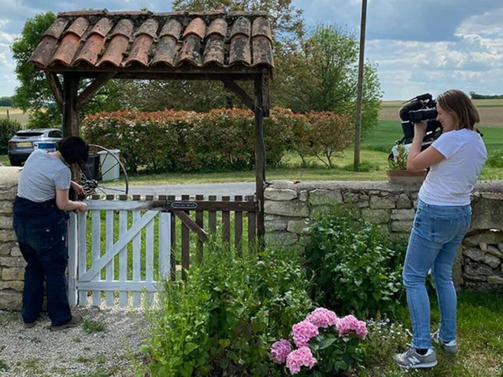 Two women in a garden. One has a video camera and the other is mending a gate