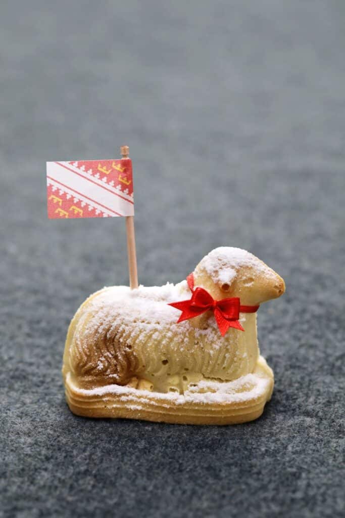 A festive Easter Lamb cake, known as Lammala, adorned with a dusting of powdered sugar and a bright red bow, holds a small flag with red and white stripes and yellow reindeers. The cake is presented against a muted grey background, emphasizing its detail and festive decoration.