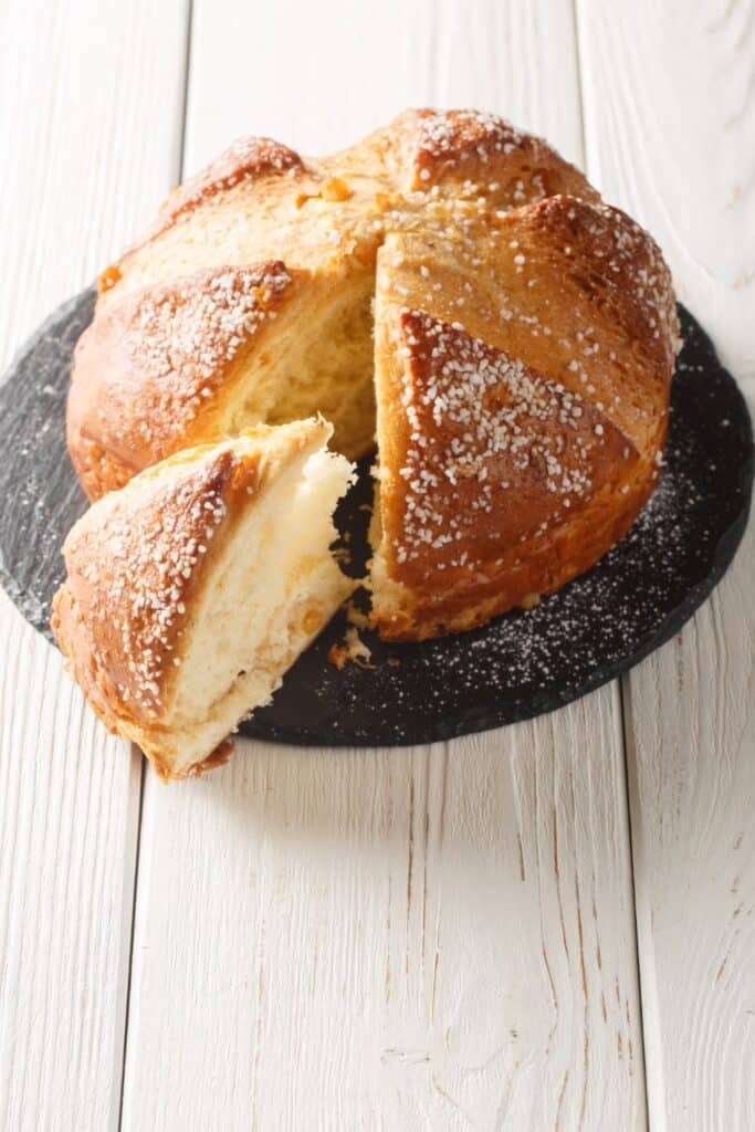 A freshly baked Easter brioche, known as 'Moana', on a slate serving plate, with one slice partially removed to reveal its soft, fluffy interior. The golden crust is sprinkled with sugar granules, adding texture and sweetness to this traditional holiday bread on a white wooden surface.