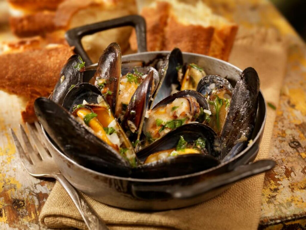 A pot of freshly cooked mussels in creamy white wine sauce, garnished with herbs, presented in a rustic cast-iron serving dish. The opened shells reveal the tender, steamed mussels inside, ready to be enjoyed. In the background, there's crusty bread on a wooden board, perfect for soaking up the flavorful sauce.