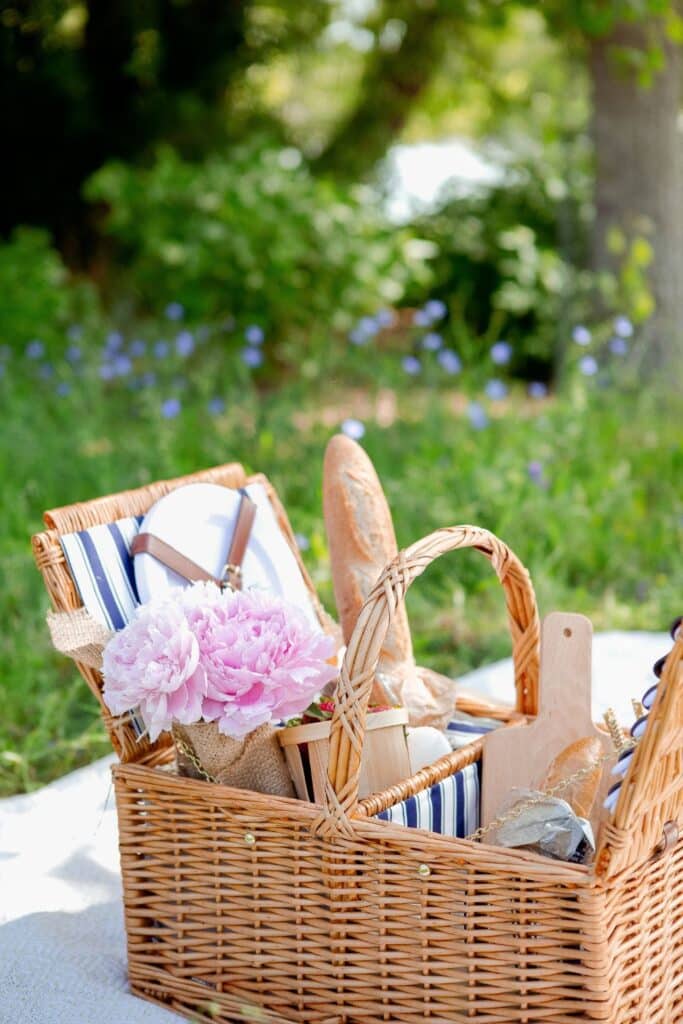 A beautifully styled wicker picnic basket filled with classic French picnic essentials, including fresh baguettes, wooden cutting boards, blue-striped napkins, and a bouquet of pink peonies. The basket sits on a soft white picnic blanket, surrounded by lush greenery and delicate wildflowers.