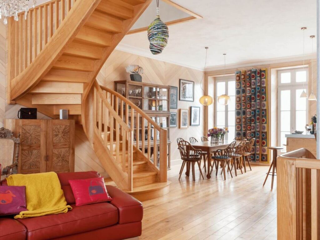 An open plan room in a house in France with a wooden floor and staircase, a sofa with cushions and a table and chairs