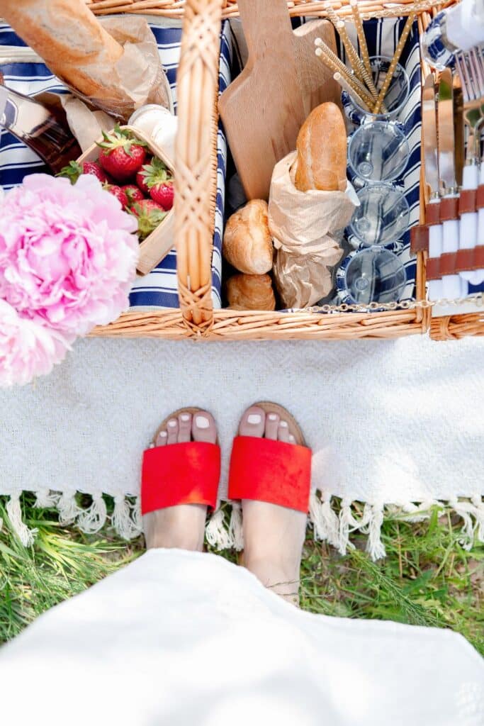A close-up of a French picnic spread with a wooden charcuterie board displaying creamy cheeses, crackers, and fresh berries. Bowls of cherries, croissants with strawberries, and a glass of rosé complete the elegant setup.