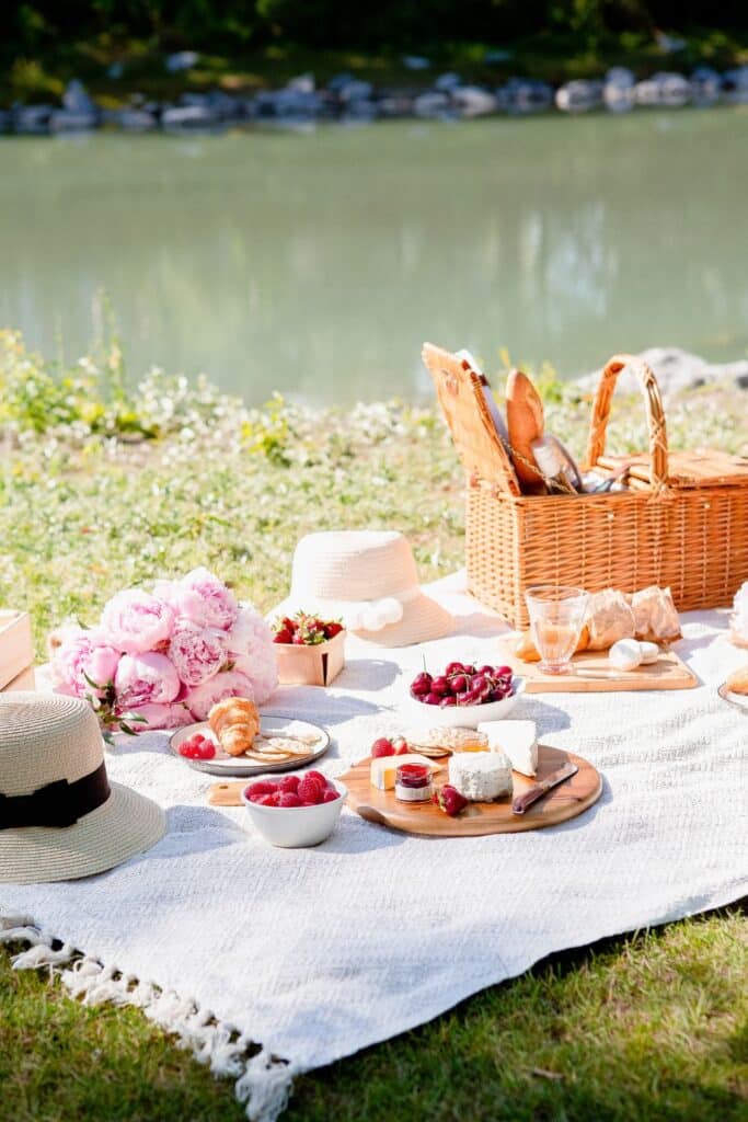 A cozy French picnic setting by a lake, featuring a soft textured blanket covered with a wicker basket, peonies, a wooden cutting board of cheese and fruit, and stylish summer hats.