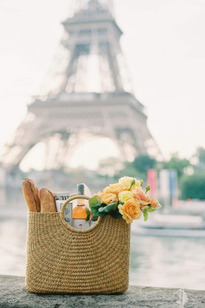 A scenic picnic scene under the trees with a rustic wooden crate holding a bottle of rosé, a red woven basket, and glasses of wine. Nearby, fresh bread, macarons, and raspberries rest on a picnic blanket.