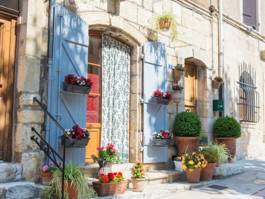 An old French building with blue shutters and an arched entranced with flower pots either side