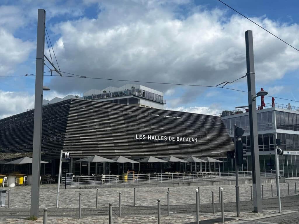 The outside of a covered market in Bordeaux with the Cite du vin in the background with a blue cloudy sky
