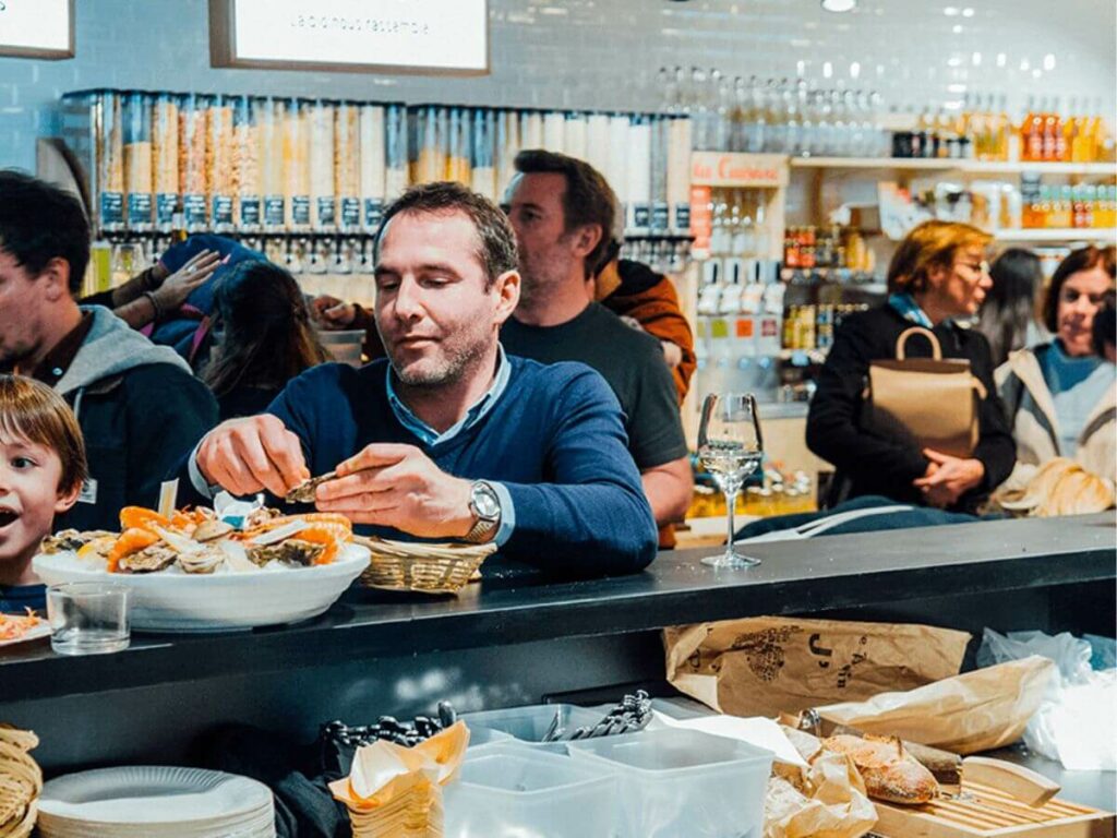 Man eating seafood at a busy cafe in a Bordeaux market with other people around