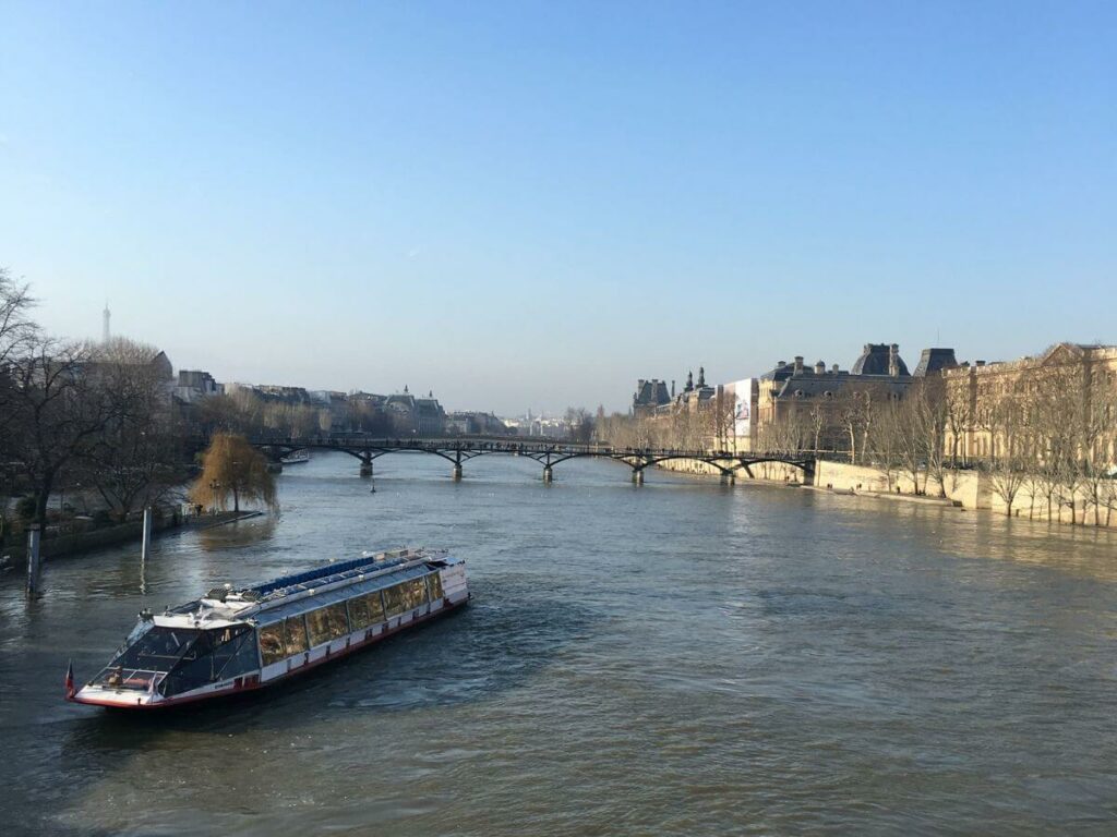 A river in France with a bridge going across and a boat sailing on it with Paris in the background