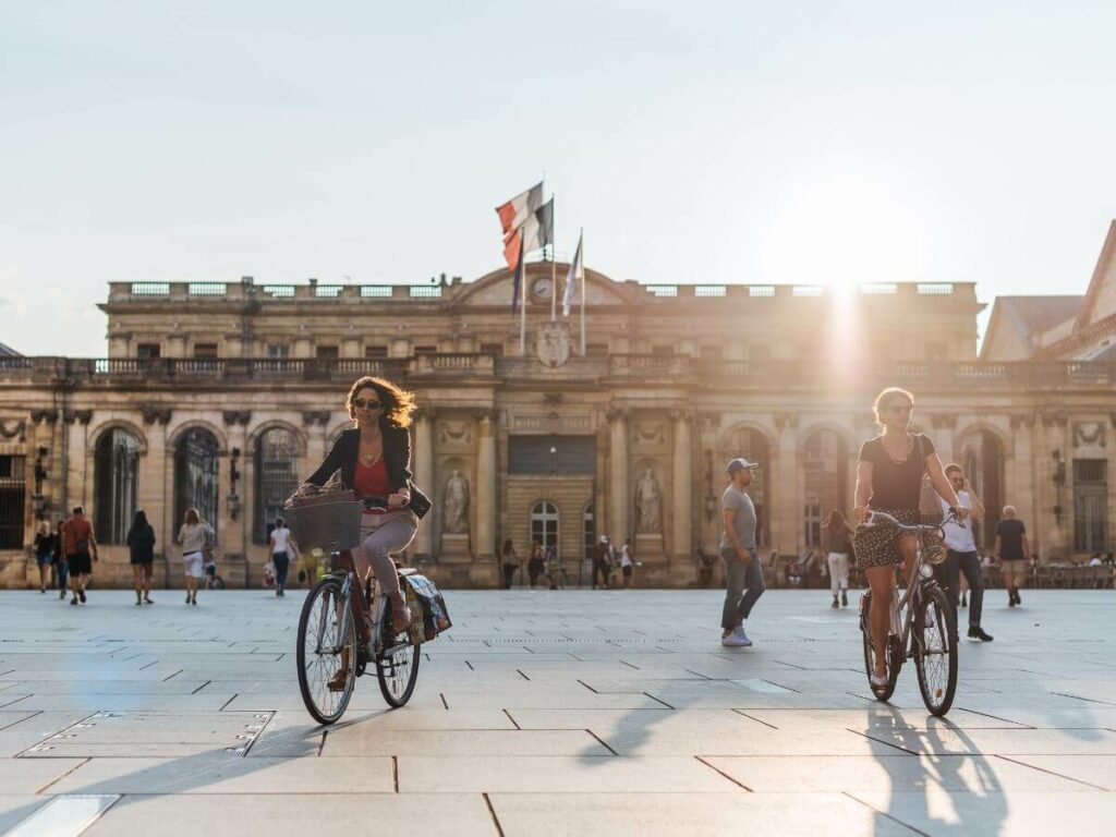 An old city building of stone in Bordeaux square with two women on bikes
