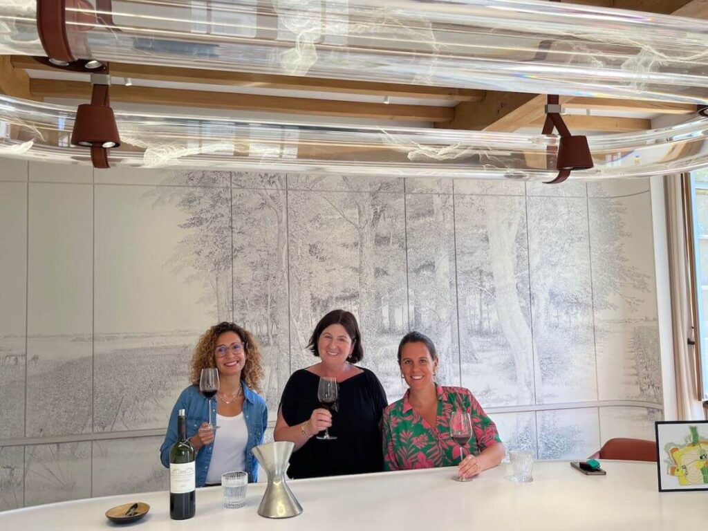 Three women with wine glasses stood against a wall of art in a tasting room in Bordeaux
