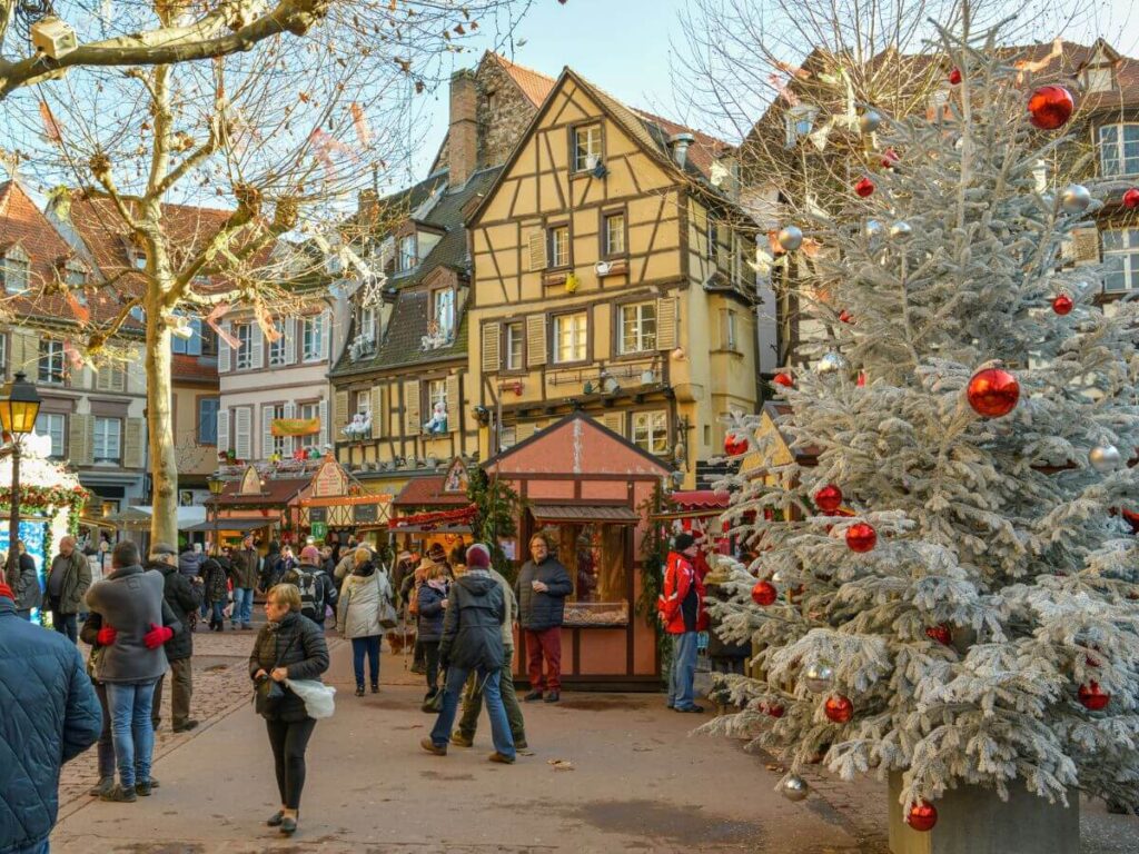 The marketplace of a Christmas Market in France with a white Xmas tree wiht red baubles, half-timbered buildings and people walking around
