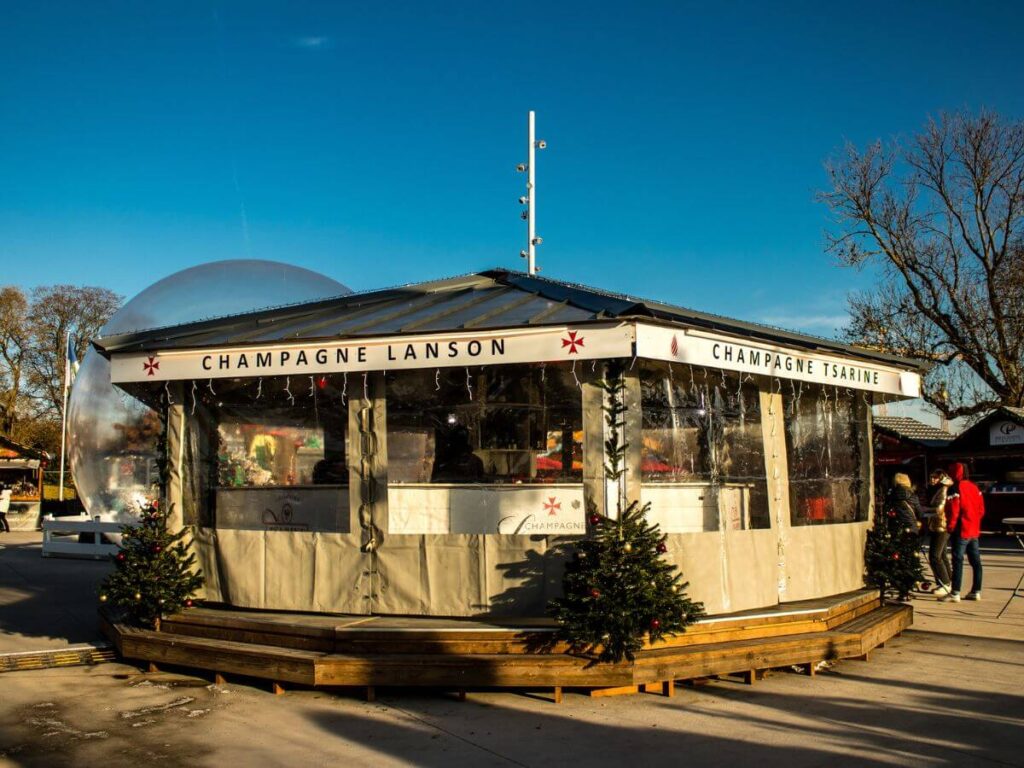 A wooden chalet at a Christmas market with Champagne Lanson written on it and Christmas Trees in front