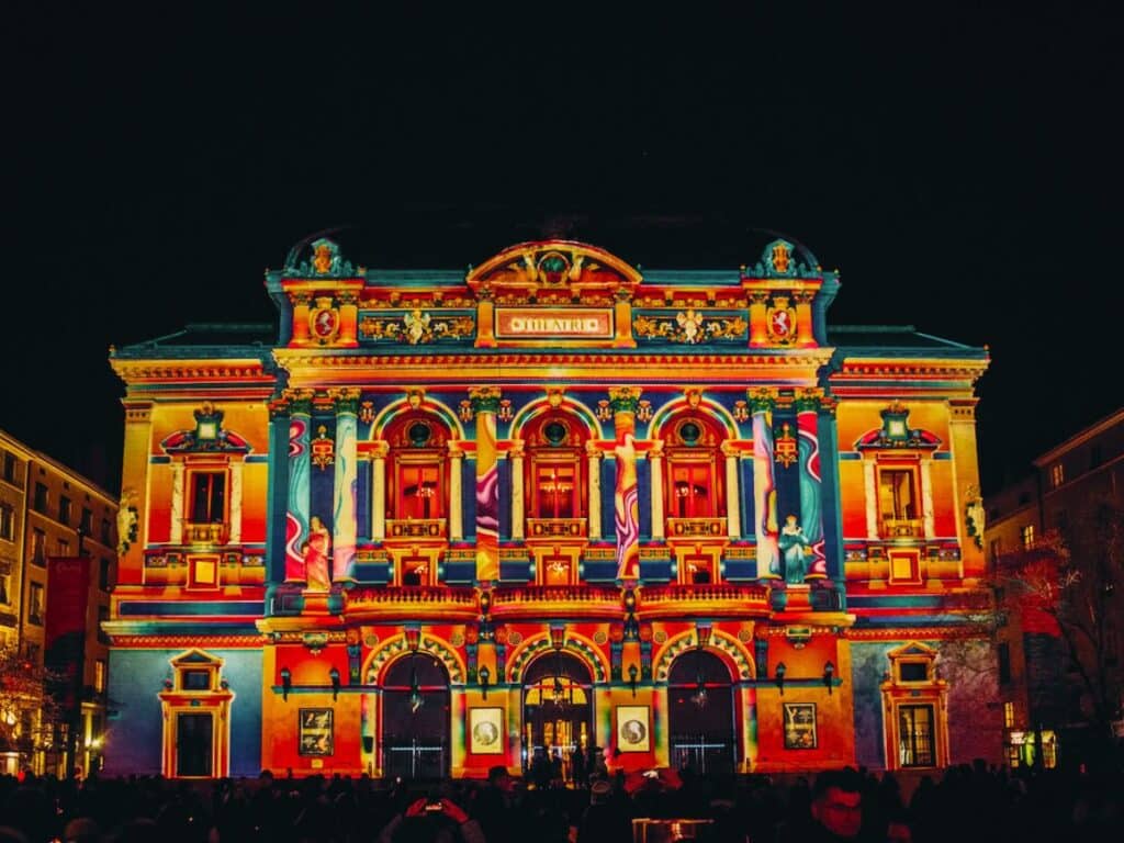 This image shows a vibrant building facade illuminated by colorful lights during what appears to be a nighttime festival, possibly the Fête des Lumières in Lyon, France. The ornate architecture of the building, likely a historic or cultural site, is lit up with striking hues of red, orange, blue, and purple, creating a festive and celebratory atmosphere with a crowd gathered in the foreground.