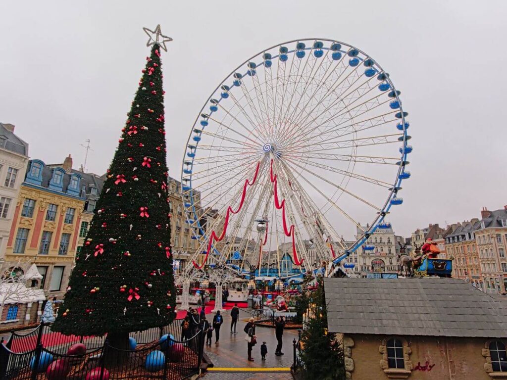 A ferris wheel and Christmas Tree set against some old buildings in France against a grey sky
